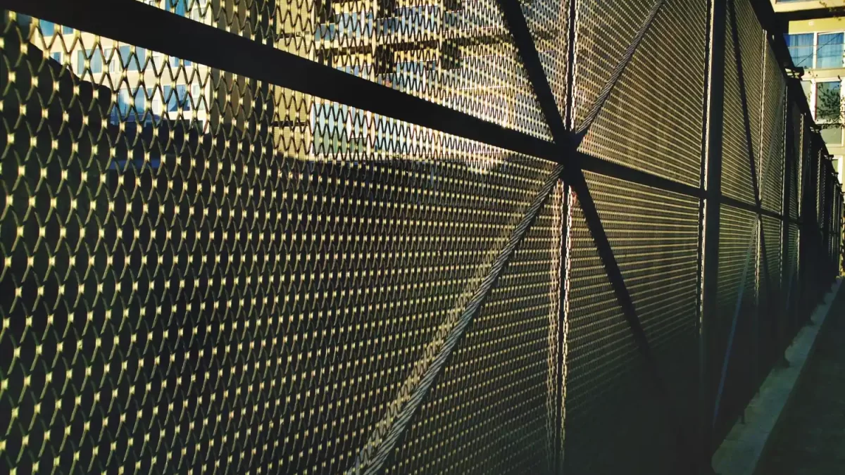 Vista lateral de uma robusta cerca de tela metálica expandida em um ambiente urbano, com edifícios ao fundo e luz natural destacando sua trama.