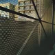 Vista lateral de uma robusta cerca de tela metálica expandida em um ambiente urbano, com edifícios ao fundo e luz natural destacando sua trama.