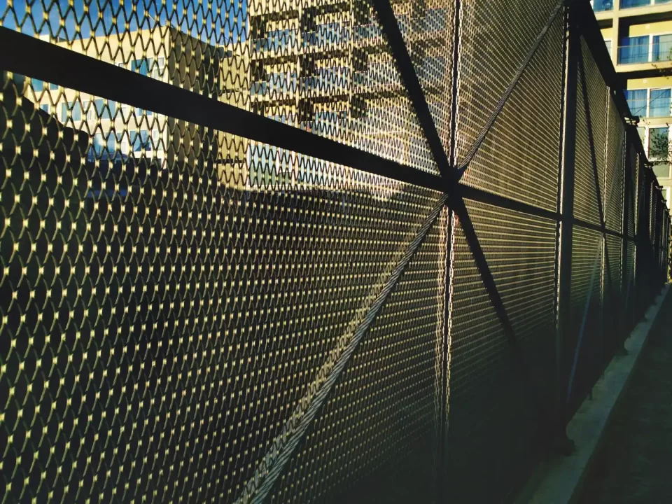 Vista lateral de uma robusta cerca de tela metálica expandida em um ambiente urbano, com edifícios ao fundo e luz natural destacando sua trama.
