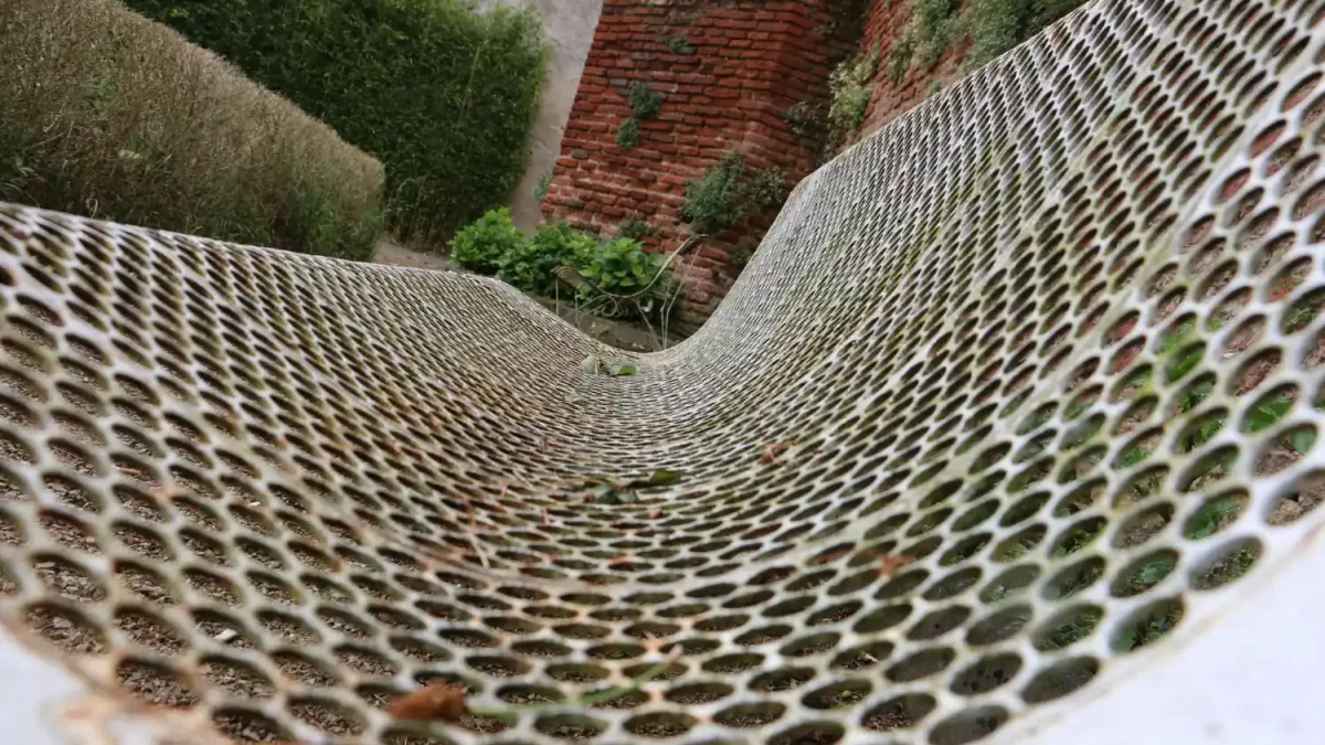 Tela expandida metálica com deformação e curvatura acentuada instalada em área externa com vegetação e muro de tijolos.