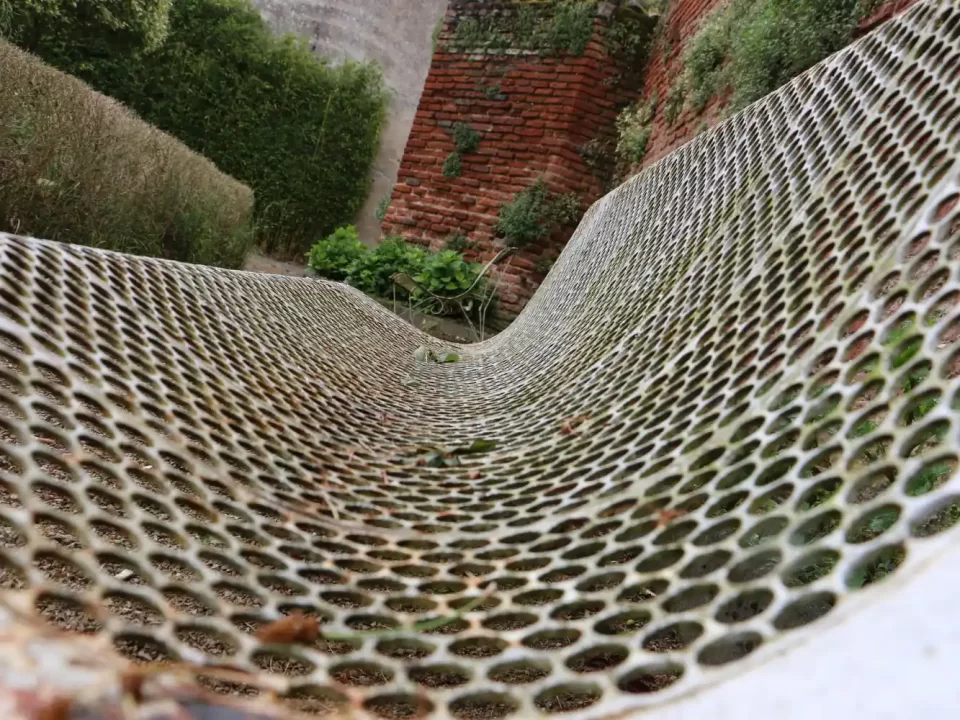 Tela expandida metálica com deformação e curvatura acentuada instalada em área externa com vegetação e muro de tijolos.